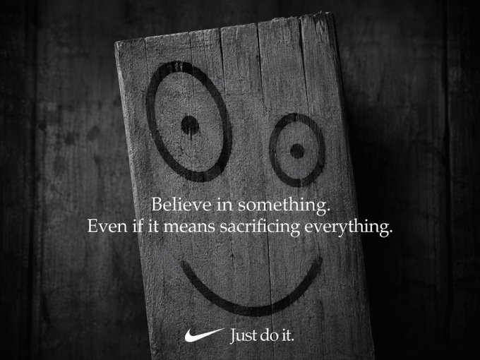 A black-and-white Nike advertisement featuring a wooden board painted with a smiley face and the text "Believe in something. Even if it means sacrificing everything. Just do it."