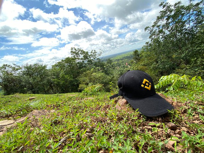 A black cap with a yellow "J" sits on a grassy hillside overlooking a lush, forested valley under a partly cloudy sky—capturing the scenic mountain view featured in the post while promoting the @JohnRealKH brand.
