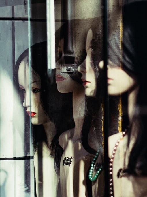 An artistic, layered reflection shows multiple overlapping images of a woman with dark hair, red lipstick, and green beaded necklaces against a dark, glassy background.