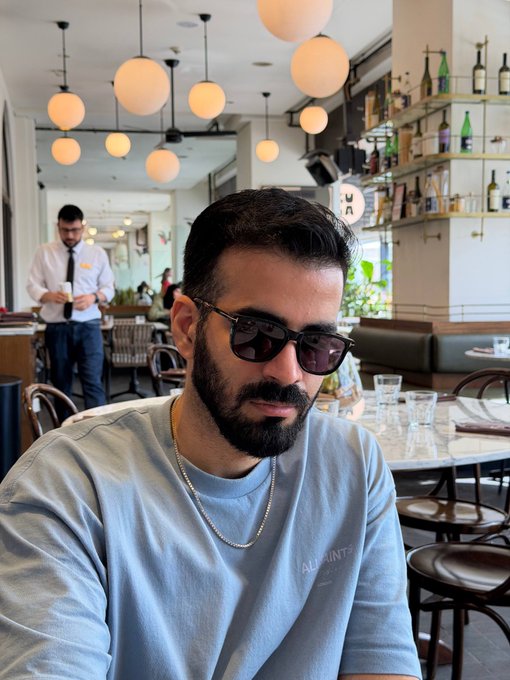 A bearded man wearing sunglasses and an "AllSaints" t-shirt poses inside a stylish restaurant/cafe with globe pendant lights and a bar stocked with bottles in the background, where a waiter in a white shirt and tie attends to a guest—capturing a relaxed weekend vibe that fits the "GM CT happy weekend" greeting.