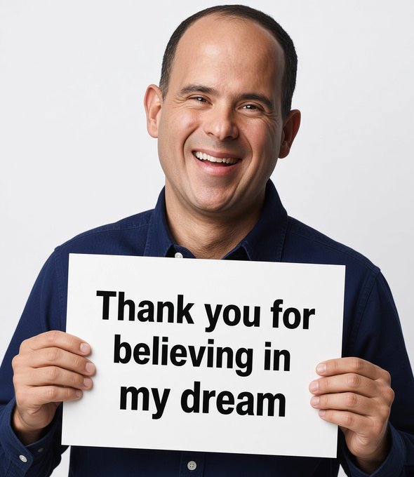 A smiling bald man in a dark blue shirt holds a white sign that reads 'Thank you for believing in my dream.'