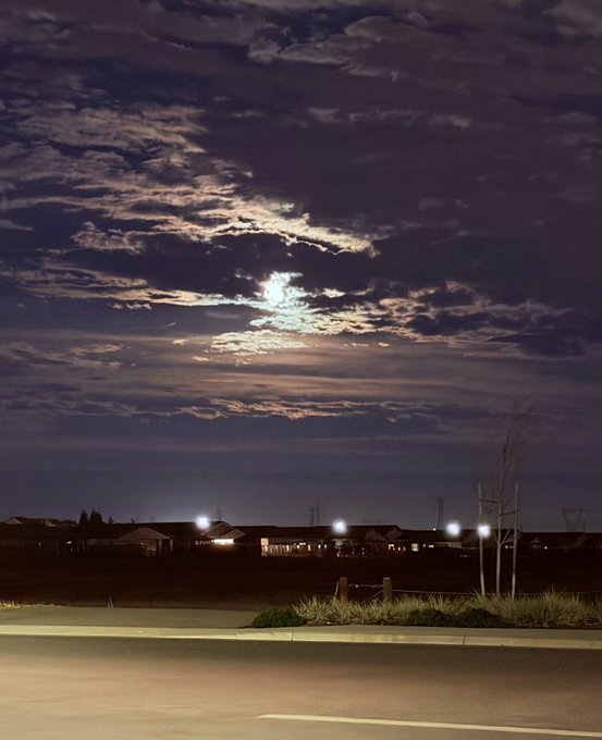 The moon shines brightly through scattered clouds over a darkened suburban neighborhood at night, with streetlights and illuminated windows dotting the horizon beneath the glowing night sky. The serene evening scene captures the quiet beauty of nighttime with the moon partially veiled by wispy clouds.