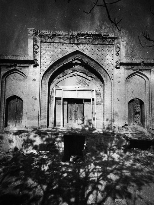A black-and-white photograph of an abandoned, dilapidated brick building with arched entryways, photographed at night under a starry sky.