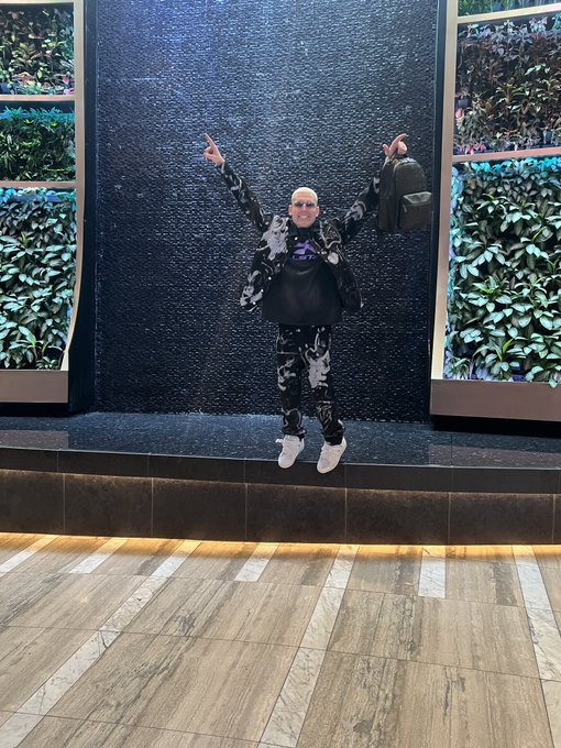 A person in a black and white floral-patterned outfit and sunglasses strikes a joyful victory pose with both arms raised in front of a textured black water wall flanked by lush green plant displays. The celebratory stance and upscale hotel-like setting visually embody the "GM to future crypto millionaires" sentiment, projecting confidence and success while ignoring the skeptics and scammers mentioned in the caption.