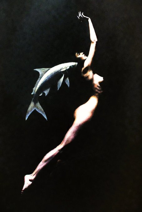 An artistic photo features a woman floating in dark water next to a large fish, with dramatic lighting highlighting her pale skin and the fish's scales.