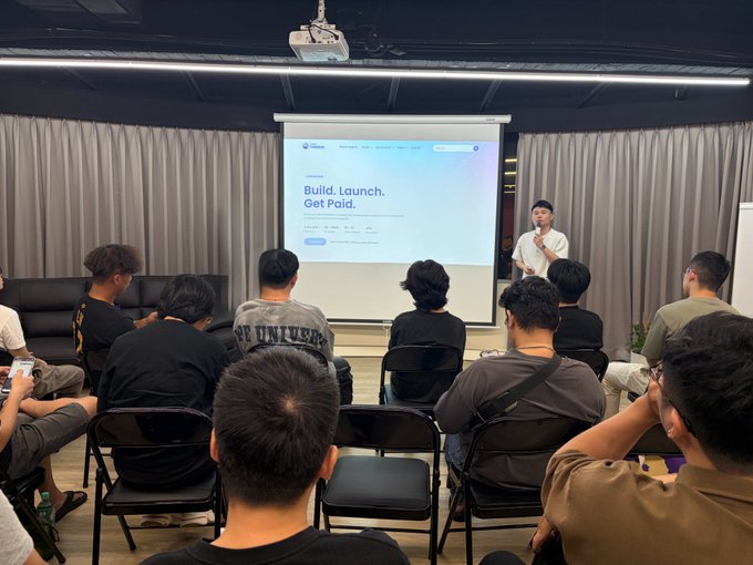 A presenter standing in front of a projection screen displaying 'Build. Launch. Get Paid.' while addressing an audience of people seated in folding chairs.