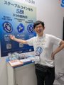 A man stands smiling in a blue t-shirt holding a Daruma doll at a SX-Blockchain and Digital Asset Summit booth, with event signage displaying "SCB 2024" visible in the background.