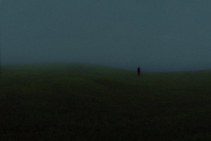 A very dark, grainy landscape photograph of a grassy hill at dusk, with a single small figure standing on the horizon line.