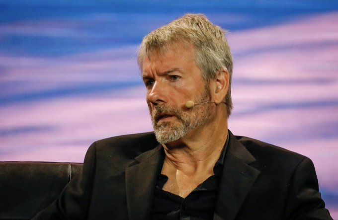 The image shows a man wearing a suit sitting in front of a blue screen with his head tilted downwards, giving him a serious expression. In the background, there are additional people present but they are blurred and not as prominent as the main subject. The context of the image suggests that it may be related to an event or gathering where Michael Saylor is speaking, as indicated by the #Bitcoin post in the caption provided.