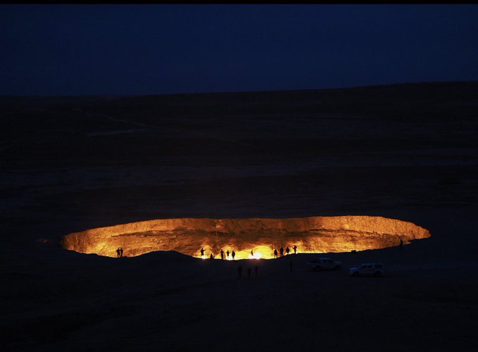 A large crater illuminated with a warm glow at night, with silhouettes of people and vehicles standing at its edge.