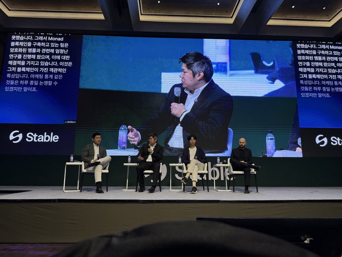 A panel of four speakers sits on stage at what appears to be the "Stable" summit, with a large screen displaying Korean translation text and a close-up of one speaker holding a microphone, reflecting the post's humor about a friendly and respectful discussion that failed to spark the anticipated "fight" between panelists.