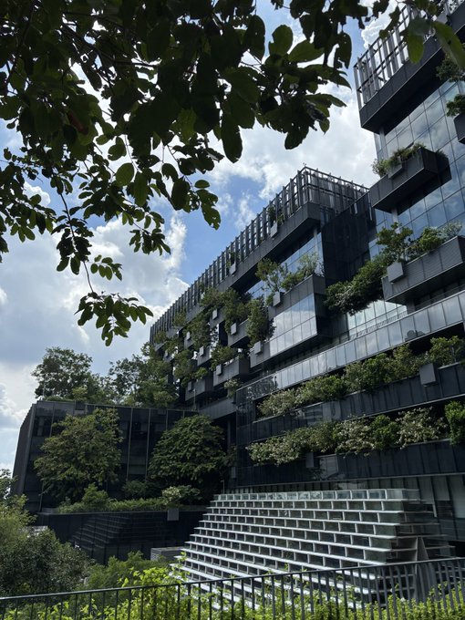 A modern, multi-story building with a glass facade is integrated with abundant greenery, featuring plants growing on terraced balconies and rooftops against a partly cloudy sky.