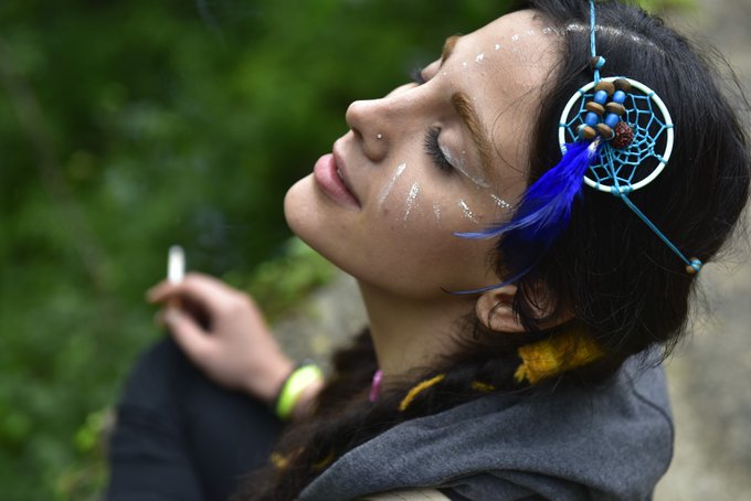 A woman with closed eyes, adorned with white face paint and a blue dreamcatcher, holds a cigarette against a blurred green background, embodying the contemplative, artistic spirit that a camera can capture but not create. The image visually echoes the post's meditation on photography as a vessel for meaning, ownership, and the photographer's unique vision—where value lies in the story and emotion behind the lens, not just the image itself.