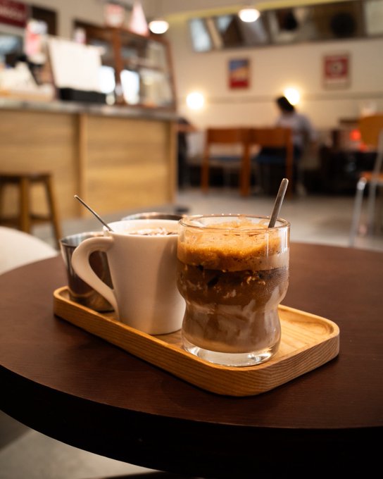 Two coffee drinks on a wooden tray at a cafe, including an iced espresso in a glass and a white ceramic mug, with a blurred interior and patron in the background.