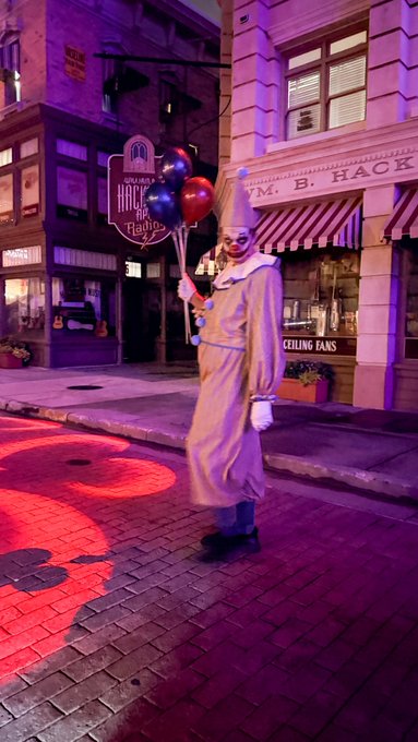 A person dressed as a clown holding balloons at night on a street with Halloween decorations and storefronts including 'M.B. HACK'.