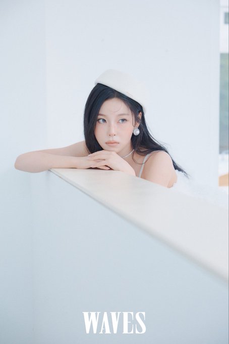 A portrait photo of a young woman with long dark hair wearing a white beret, resting her arms on a white surface, with the word 'WAVES' at the bottom.