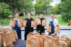 Four people in aprons stand behind a long table filled with brown paper bags, posing with bags in hand at what appears to be a food distribution or charity event outdoors.