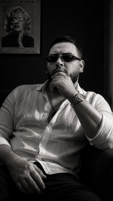 Black and white portrait of a bearded man wearing sunglasses and a light button-up shirt, posed with his hand on his chin, with a Marilyn Monroe poster visible on the wall behind him.