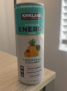 A 12 fl oz (355 mL) can of Kirkland Signature Sparkling Tropical Energy Drink sits on a wooden surface, featuring 200 mg caffeine, 7 essential vitamins, and zero sugar. The image supports the post's theme by showcasing a tropical-flavored energy drink as a Costco-brand alternative to Celsius.