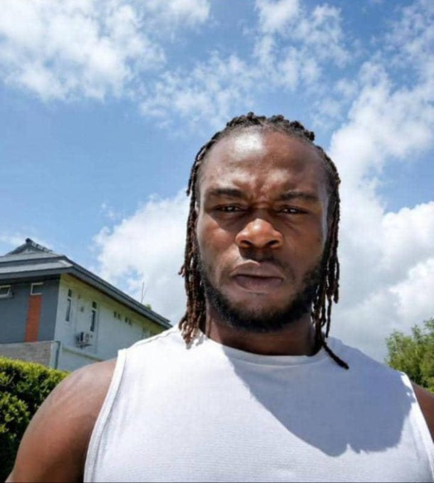 A selfie of a muscular man with dreadlocks wearing a white tank top outdoors, with a blue house and cloudy sky in the background.