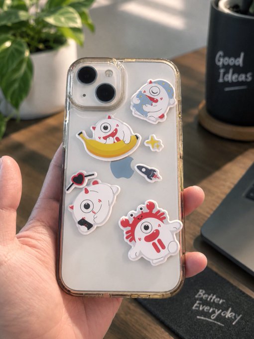 A hand holding a clear smartphone case decorated with various cartoonish creature charms, positioned on a desk next to a mug reading 'Good Ideas'.