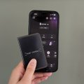 A hand holds a black Ledger hardware wallet in front of a smartphone displaying what appears to be a crypto portfolio app with a balance of "1 600 545.21," showcasing the essential hardware-software combo for securely managing cryptocurrency holdings.