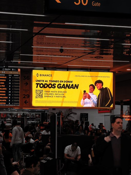 The image shows a Binance advertisement in Spanish displayed on a digital billboard at what appears to be an airport or transit hub, reading "BINANCE - ÚNETE AL TORNEO EN DÓNDE TODOS GANAN - GANA HASTA $100,000 DÓLARES. INICIA EN: BINANCE Y PARTICIPA," featuring two people with smartphones, directly promoting the Binance tournament referenced in the post.