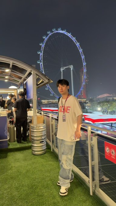 A young man wearing a white 'SLOPE' t-shirt and blue jeans stands on a balcony at night, with a large illuminated Ferris wheel and city skyline in the background.