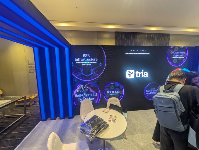 An indoor trade show or convention booth displays a large illuminated wall with the logo 'tria' and promotional graphics for 'B2B Infrastructure,' 'Self-Custodial by Design,' '600,000+ supported networks,' 'INVITE ONLY,' 'Self-custody Multi-chain Infrastructure,' '200+ networks supported on one instance,' and 'Trusted global compliance,' with a person wearing a gray backpack standing nearby.
