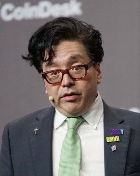 The image features a man wearing glasses, a suit, and a green tie with multiple pins attached to his jacket. The man appears to be giving a speech or presentation, as indicated by the presence of a podium behind him. In the background, there is a sign that reads "CoinDesk". The text on this sign suggests that the image might be related to cryptocurrency news or events.

The main focus of the image is the man in the suit and tie, who seems to be delivering a speech or presentation. The presence of multiple pins on his jacket indicates that he may have some affiliation with an organization or cause related to cryptocurrency.