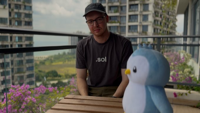 A man wearing a black cap, glasses, and a grey t-shirt with '.sol' printed on it sits at a wooden table on a balcony, with a cartoonish blue bird figurine beside him.