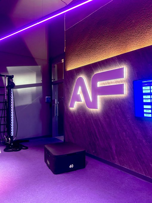 An indoor room bathed in purple lighting features a large illuminated 'AF' sign on the wall, a digital fitness display, and a plyometric box labeled '40'.