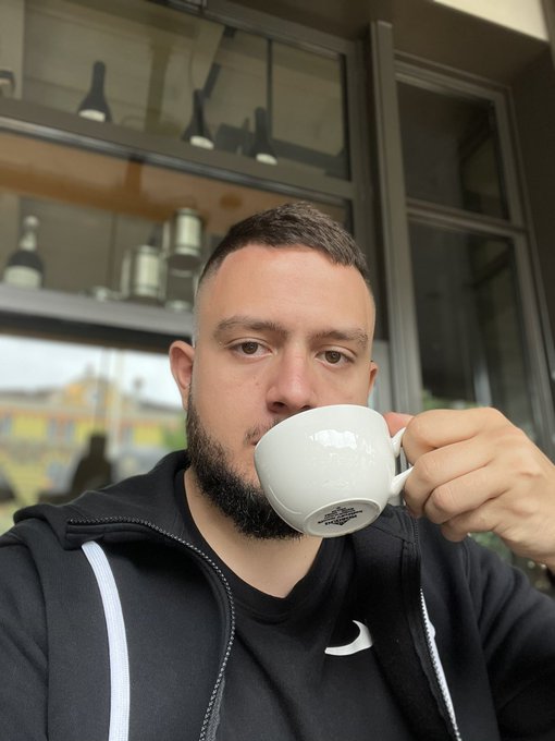 A bearded man in a black Nike hoodie drinks from a white ceramic mug, photographed inside what appears to be a cafe.