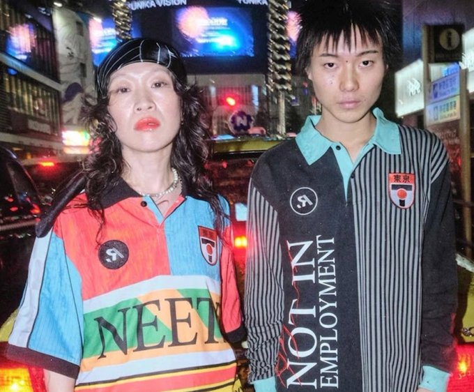 The image features two young Asian women standing together against a city street backdrop at night. They appear to be posing for a photo or possibly participating in an event related to the term "not in employment." The context of their presence suggests that they might be attending a themed gathering, protest, or social cause. Their attire and expressions convey a sense of unity and purpose as they stand together under the city lights.
