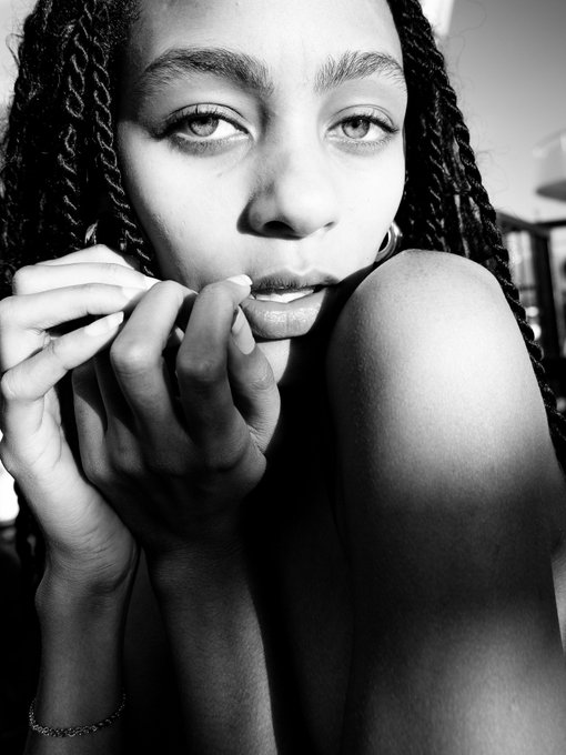 A black-and-white close-up portrait of a young woman with braided hair resting her chin on her hands.