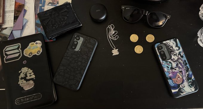 An overhead view of various personal items on a dark surface, including wallets with stickers, two smartphones, sunglasses, an AirPods case, coins, and a small white figurine.