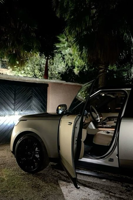A photograph of a light-colored luxury SUV with its driver-side door open at night, parked outside near a wooden gate and tropical plants.
