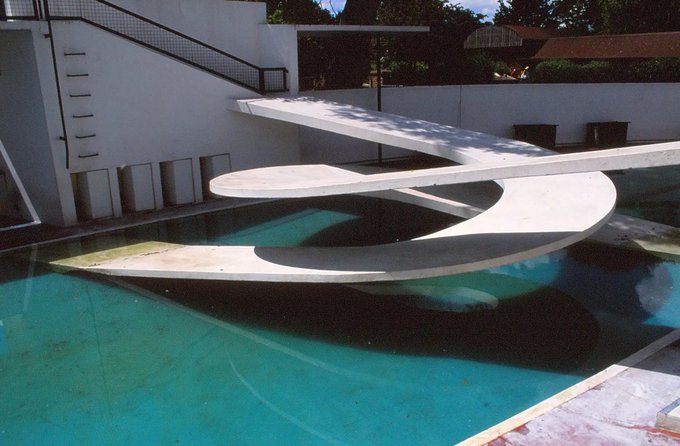 A modernist architectural installation features a sculptural, looping white concrete structure suspended over a turquoise swimming pool, set against minimalist geometric walls and surrounding greenery. No text or numbers are visible in the image.