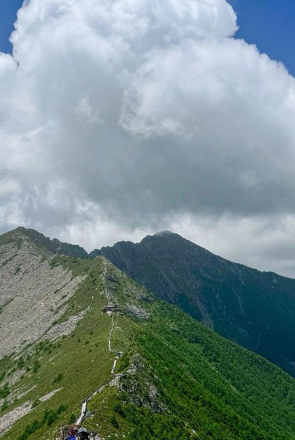 A scenic mountain landscape featuring a lush green ridge with a winding path, under a sky filled with large, billowing white clouds.