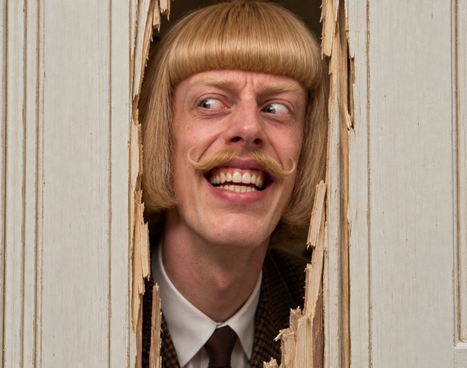 A man with a blonde bob haircut, pale skin, and a curled mustache smiles widely while peering through a hole broken in a white wooden door or wall.