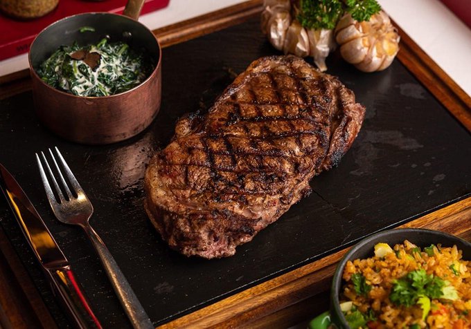 A perfectly grilled ribeye steak with attractive char marks is served on a slate platter, accompanied by creamy spinach in a copper bowl, roasted garlic, and a side of seasoned rice. The hearty, restaurant-quality presentation perfectly complements the inviting invitation to relax and enjoy a delicious dinner together.