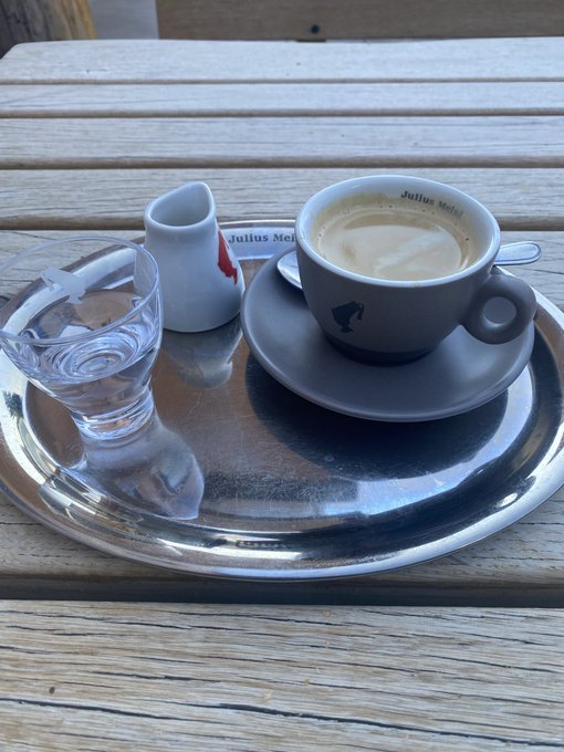 A cup of Julius Meinl coffee sits on a silver tray alongside a glass of water and a small creamer pitcher, perfectly capturing the essence of a simple yet elegant breakfast. The branded cup and cozy setting complement the weekend morning vibe.