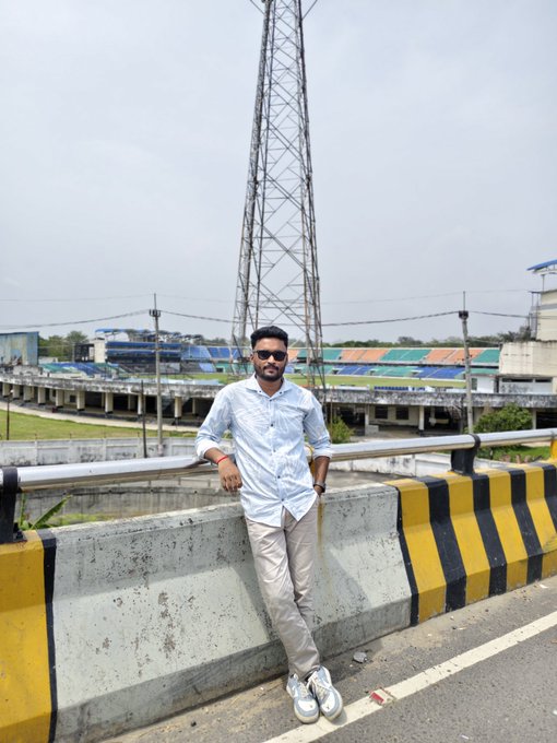 The image features a man standing next to a yellow and black striped barrier or guardrail while wearing a white shirt and khaki pants. The man appears to be posing for a photo, possibly at an event or gathering. In the background, there is a building with a tower on top of it, which could be a landmark or a significant structure in the area. Additionally, there are several cars parked nearby, suggesting that this location might be a busy street or parking lot. The image also contains text elements such as "Good evening X legend", "@useTria", and "@base".