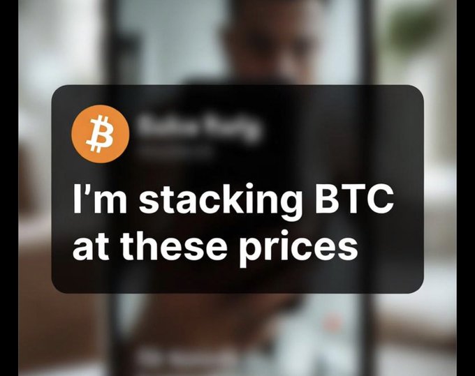 The image shows a blurred background of a person holding a smartphone, overlaid with a dark text box displaying the Bitcoin logo and the message "I'm stacking BTC at these prices." This directly reinforces the post's question about buying Bitcoin at current prices, visually emphasizing the strategy of accumulating BTC regardless of market conditions.