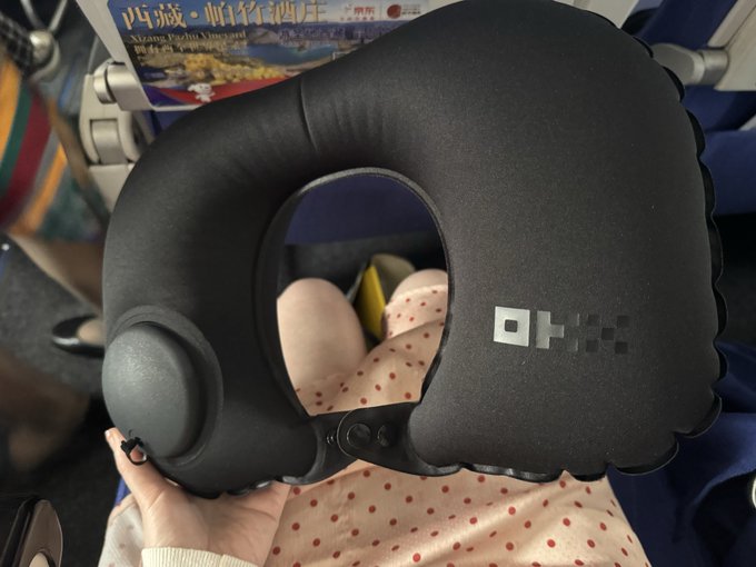 A person holds a black inflatable neck pillow branded with the "OKX" logo, shown in an airplane cabin seat with a magazine in the background. The post praises the OKX-branded inflatable neck cushion as a compact, travel-friendly essential for flights, and the image directly displays the product being recommended.