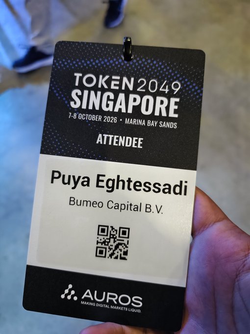 A hand holds a black attendee badge for 'TOKEN2049 SINGAPORE' dated 7-8 October 2026 at Marina Bay Sands, issued by AUROS to Puya Eghtessadi from Bumeo Capital B.V.