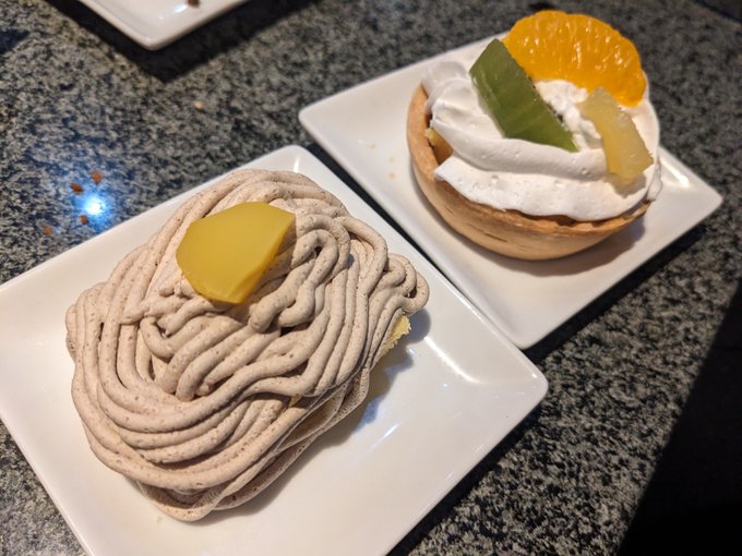 Two desserts on square white plates: a chestnut Mont Blanc-style cake with a yellow jelly topping and a tart topped with whipped cream, an orange slice, and green jelly, placed on a speckled granite surface.