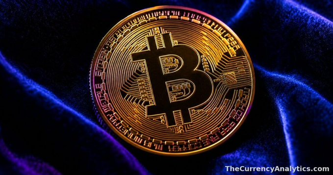 A close-up photograph of a gold Bitcoin coin resting on blue velvet fabric, with the watermark 'TheCurrencyAnalytics.com' in the bottom right corner.
