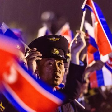 A man in a military or police officer's cap saluting while surrounded by people waving Thai flags, set against a dark nighttime background.