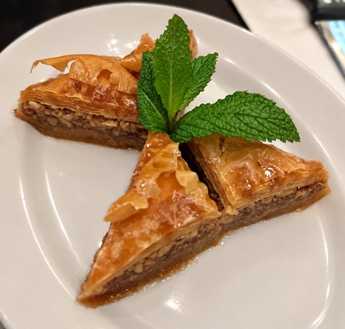 Three triangular slices of baklava, a layered pastry made with phyllo dough and nuts, arranged on a white plate and garnished with a fresh sprig of mint.
